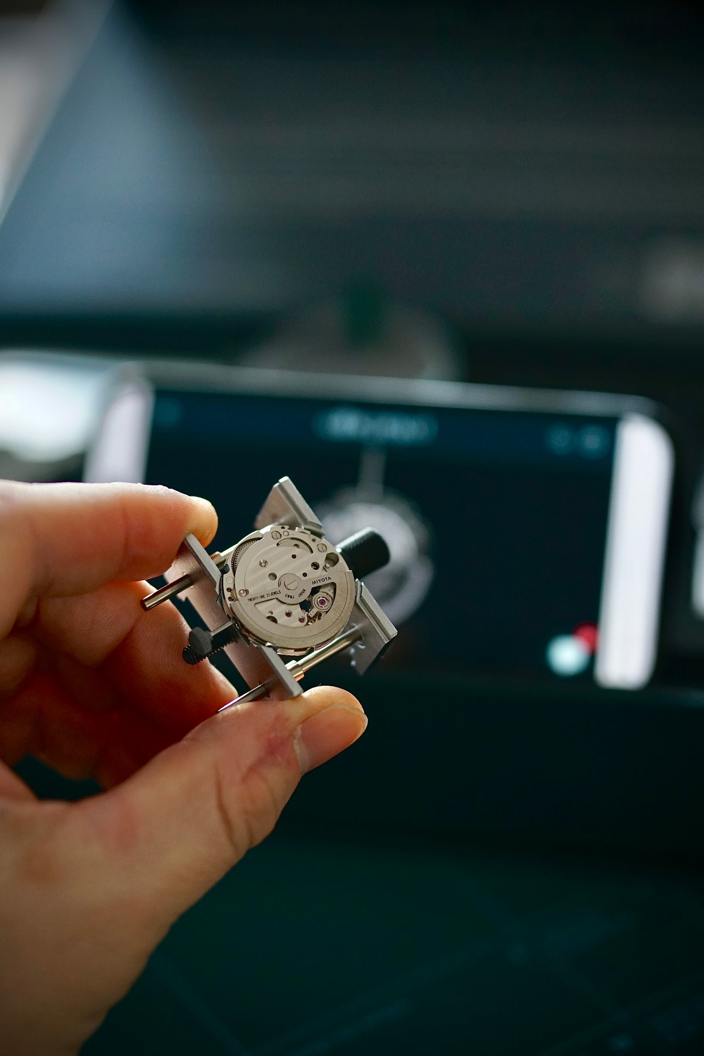 Hand holding the movement in its holder while a guided video lesson plays on the phone stand